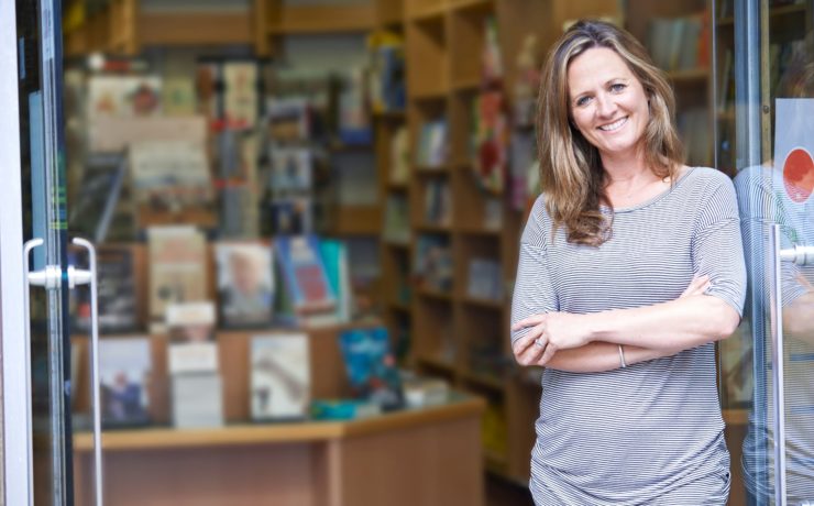 Female bookstore owner poses smiling with arms crossed in entry to her local bookstore.