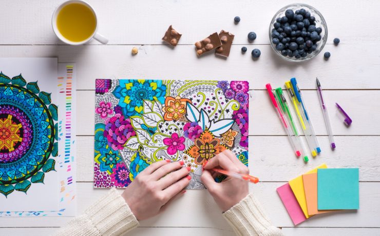 Woman using gel pens to color an adult coloring sheet surrounded by a cup of tea, blueberries pens and sticky notes.