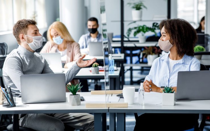 Coworkers in an office having a conversation with masks on through a space divider to prevent COVID.