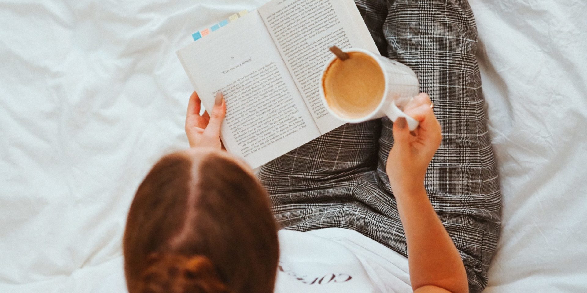 Woman sitting on a white bed reading a book and drinking coffee.