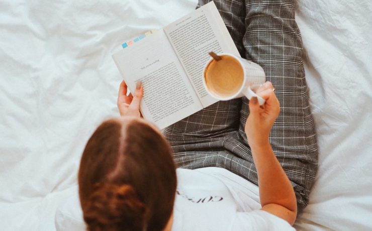Woman sitting on a white bed reading a book and drinking coffee.